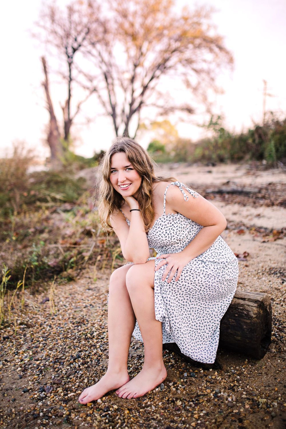 Smiling young woman in white floral dress sitting barefoot on log on rocky beach at sunset.