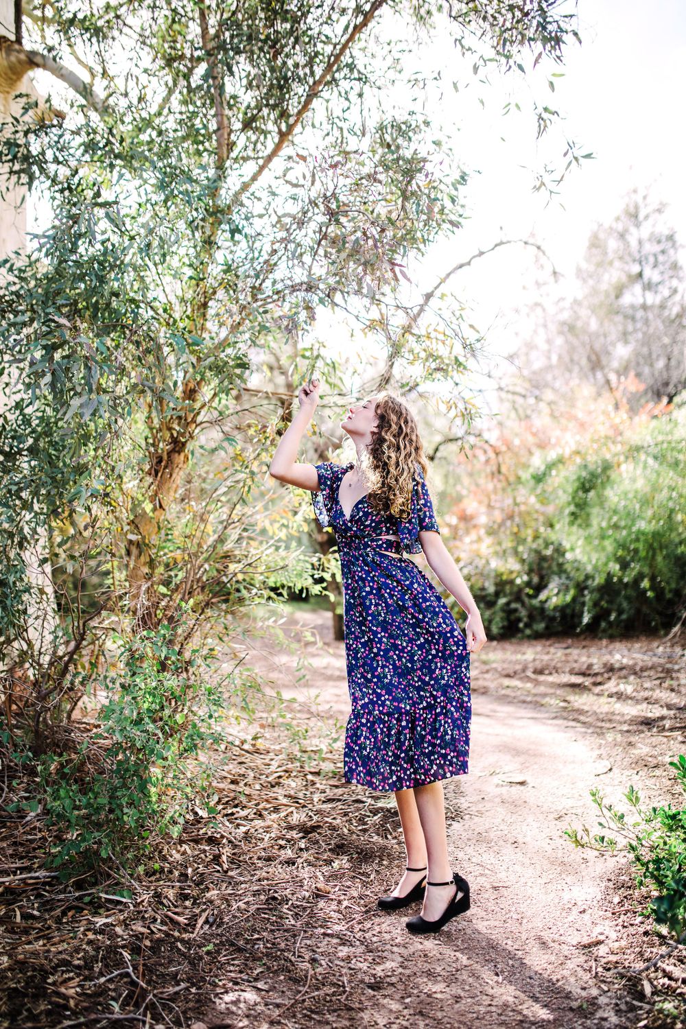 Young woman in navy floral dress reaching up to touch tree branches on a path.