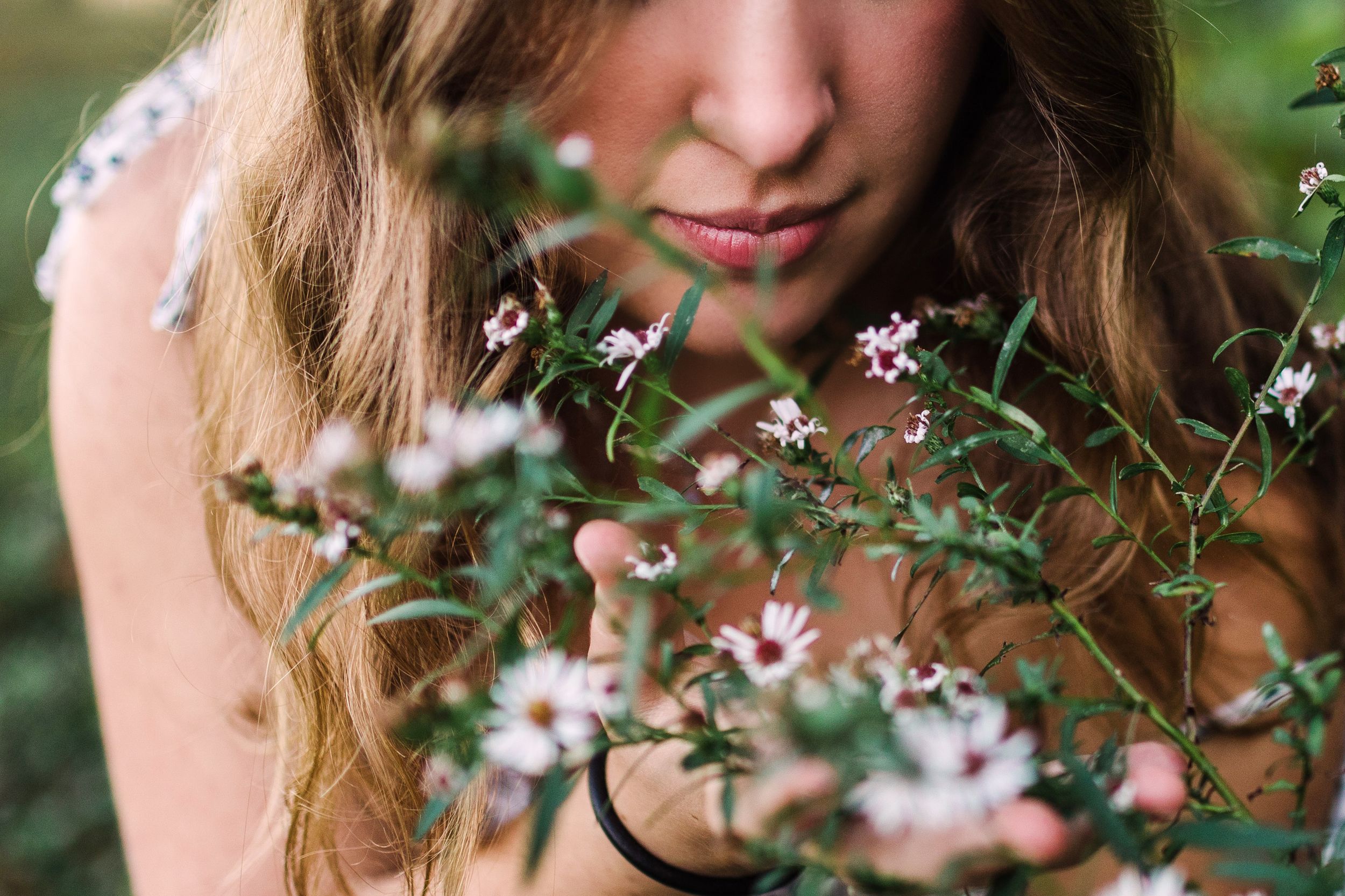 Close-up of young woman with wavy hair smelling delicate small pink wildflowers outdoors.