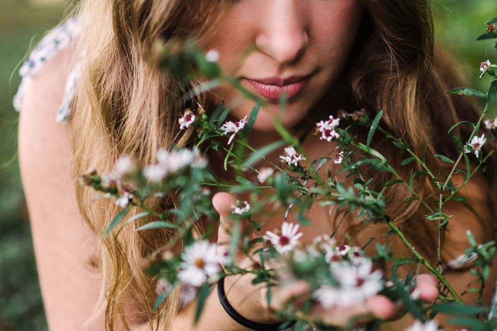 Close-up of young woman with wavy hair smelling delicate small pink wildflowers outdoors.