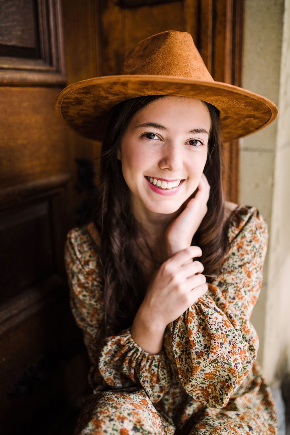 Close-up portrait of smiling young woman in brown hat and floral dress by wooden door.