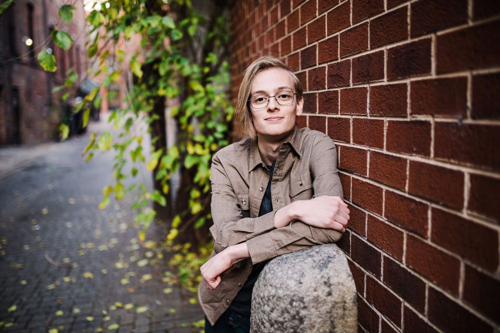 Young person with glasses sitting on stone ledge leaning against red brick wall in autumn alley.
