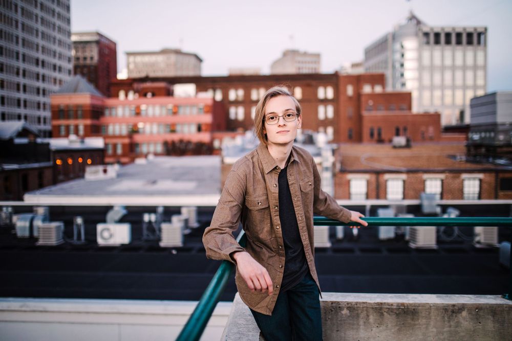 Young person in brown jacket leaning on rooftop railing with urban cityscape at sunset.