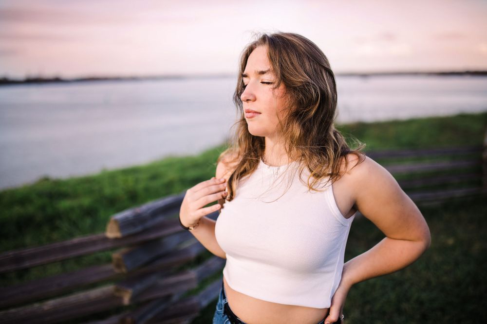 Young woman in white crop top poses outdoors near water at sunset with eyes closed.