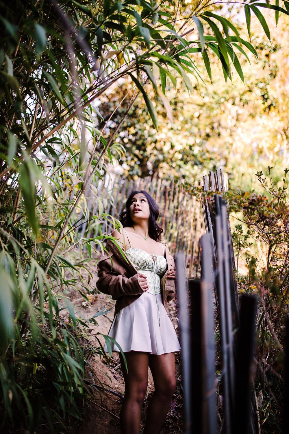 Young woman in white dress and brown jacket gazes upward in a serene bamboo garden setting.
