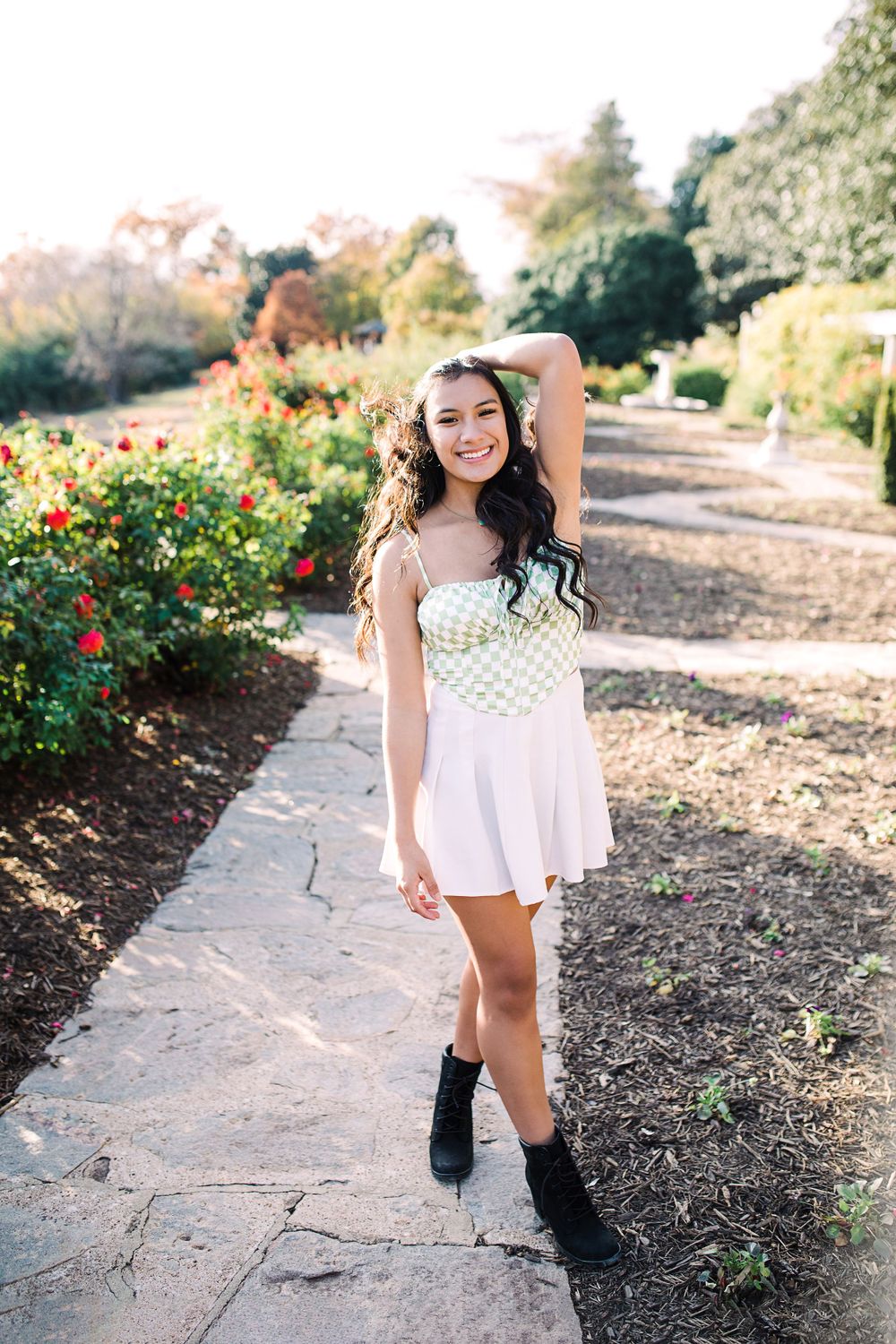 Young woman in green checkered top and white skirt smiles while posing in a vibrant rose garden.