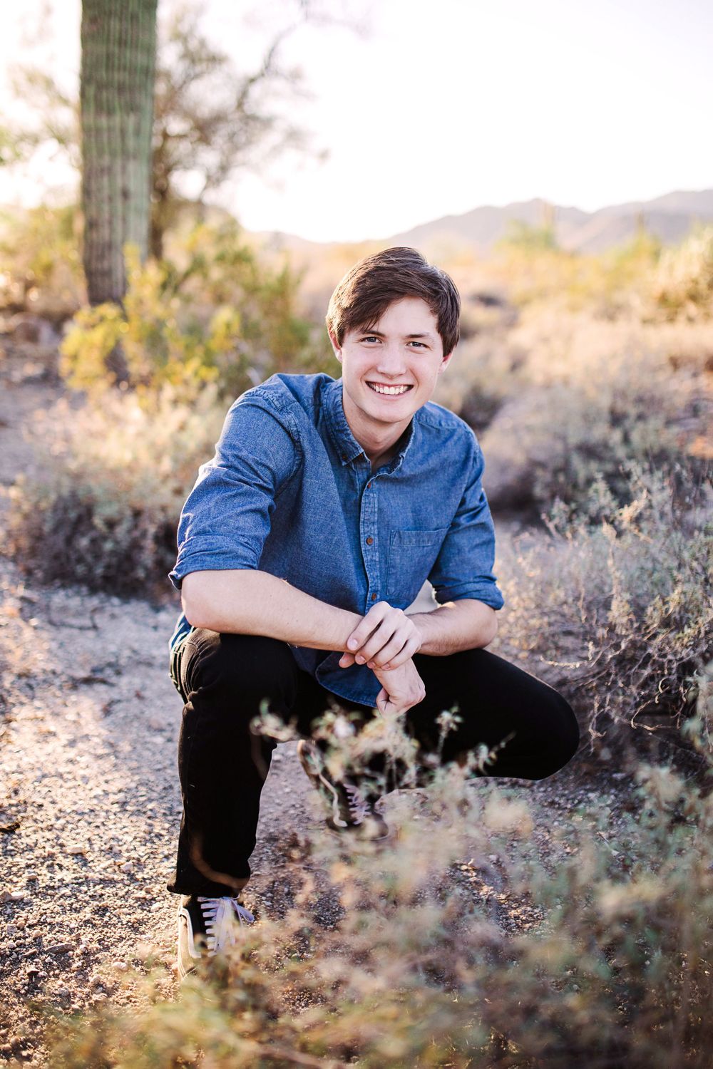 Smiling young man in denim shirt crouching in Arizona desert landscape with saguaro cacti at golden hour.