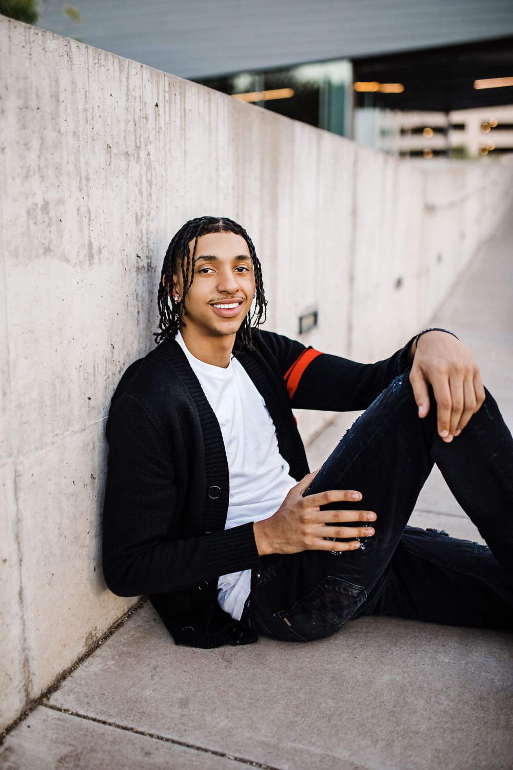Smiling young man with braids in black cardigan sits relaxed against concrete wall outdoors.