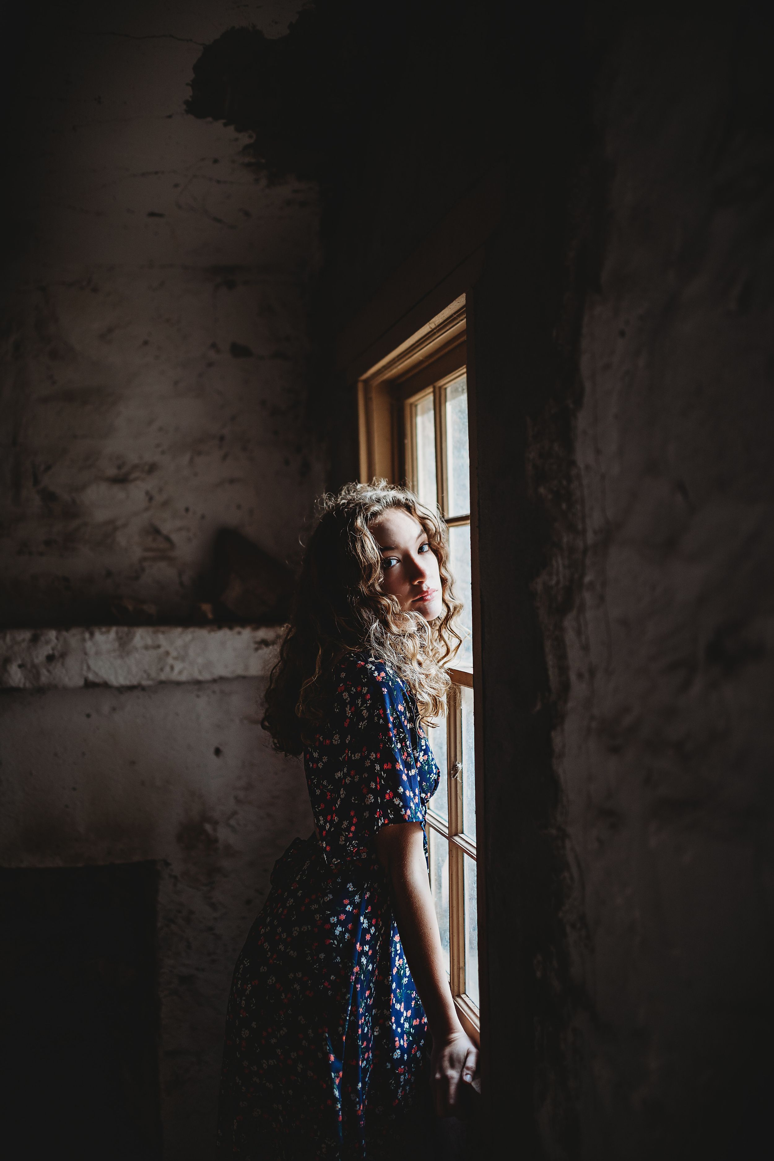 Blonde curly-haired woman in blue floral dress standing by a rustic window in a dimly lit stone room.
