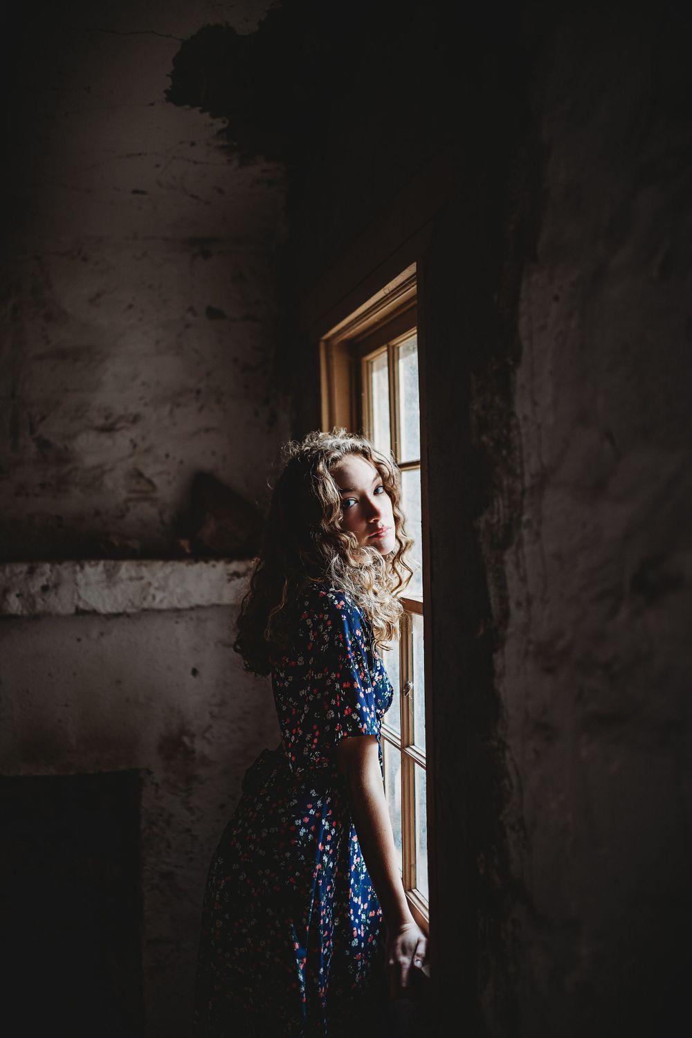 Blonde curly-haired woman in blue floral dress standing by a rustic window in a dimly lit stone room.