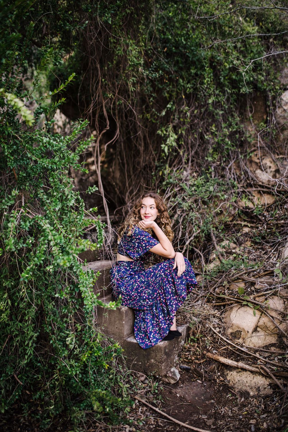 Woman in blue floral dress sitting on stone steps surrounded by lush green vines and foliage.