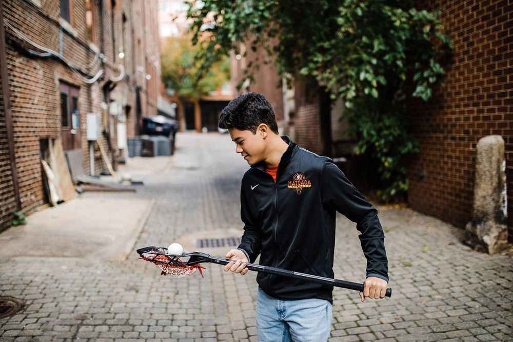 Teen boy in black Nike jacket walking in brick alley holding lacrosse stick.