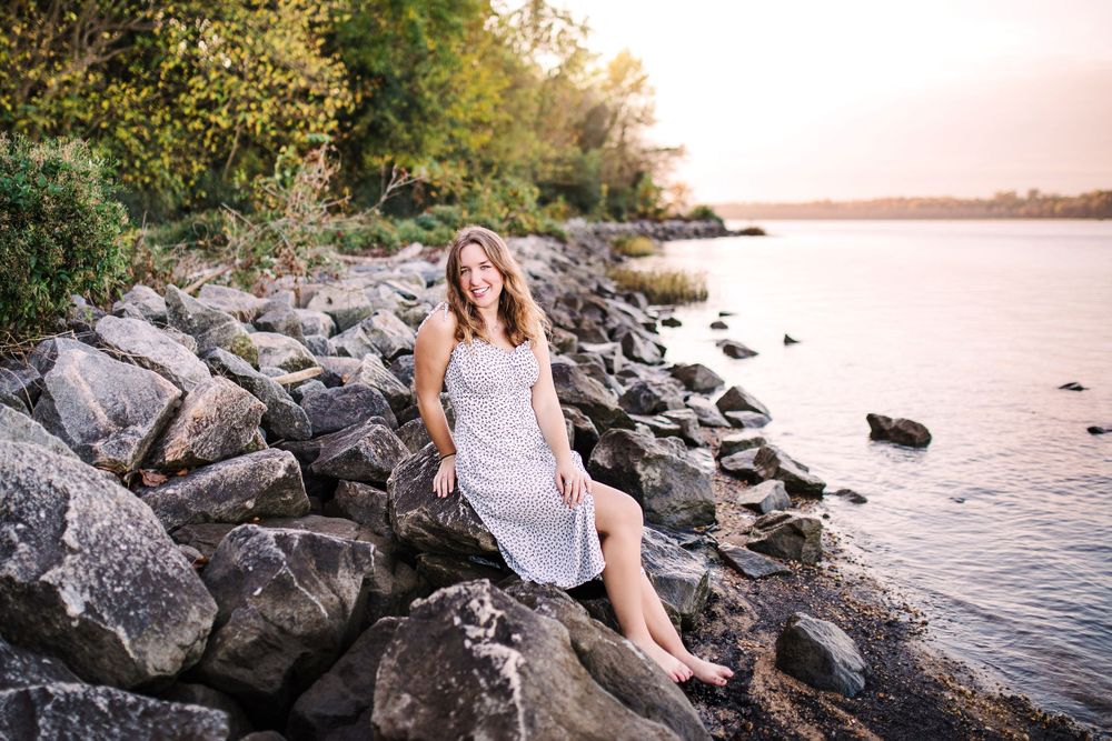 Smiling young woman in white lace dress sitting on rocky shoreline at golden hour sunset.