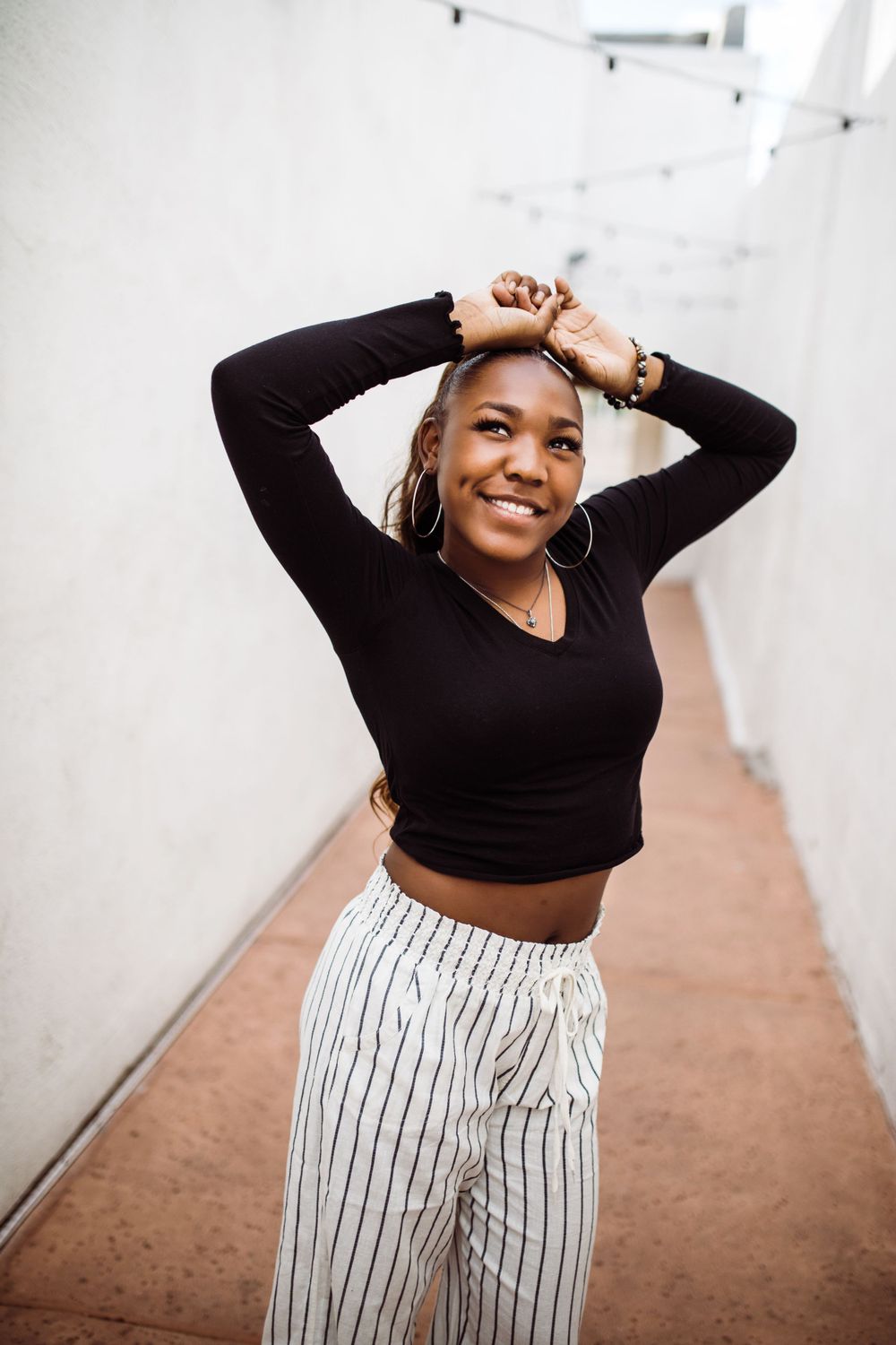 Young Black woman in black crop top and striped pants poses with hands in hair in narrow corridor.