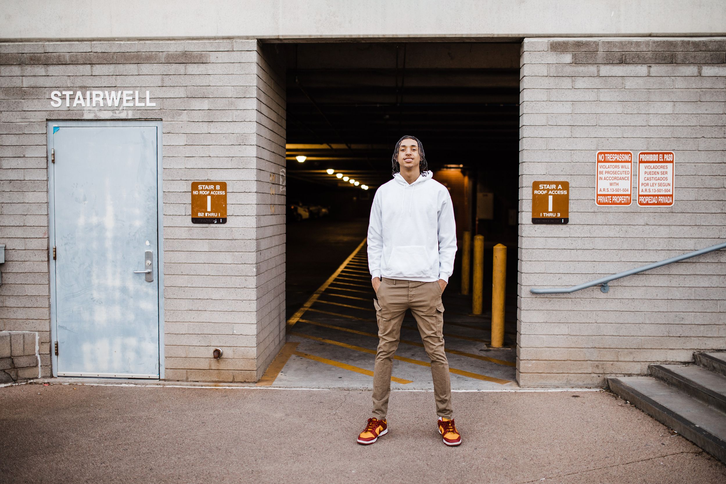 Young man in white hoodie stands outside parking garage stairwell entrance on urban street.