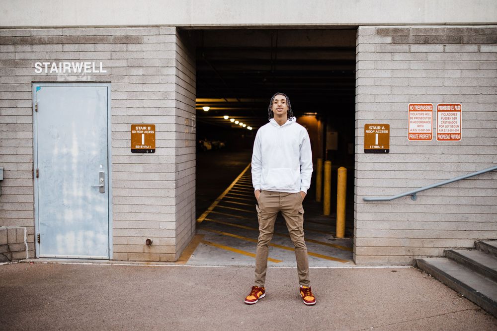 Young man in white hoodie stands outside parking garage stairwell entrance on urban street.