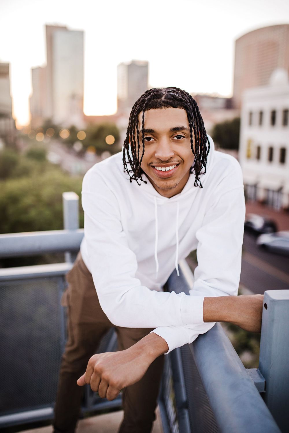 Smiling young man with dreadlocks in white hoodie leans on railing with city skyline behind him.