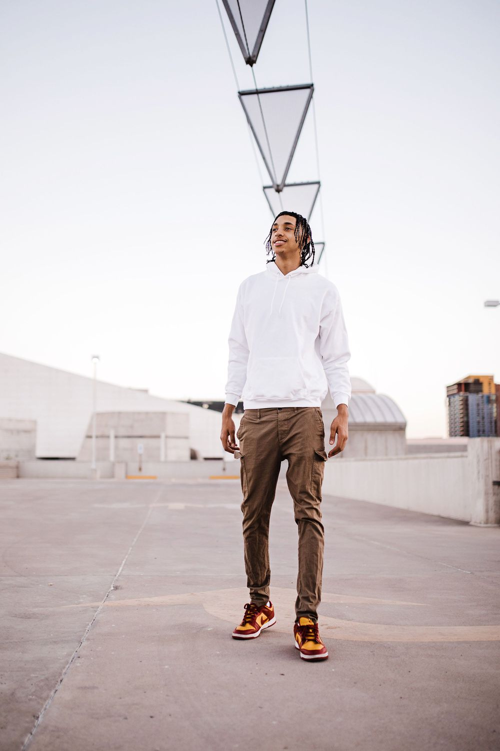 Young man in white hoodie and brown cargo pants stands on rooftop wearing orange Jordan sneakers.