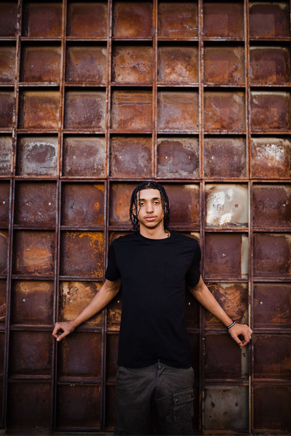 Young man with dreadlocks in black t-shirt leans against rustic metal grid wall backdrop.