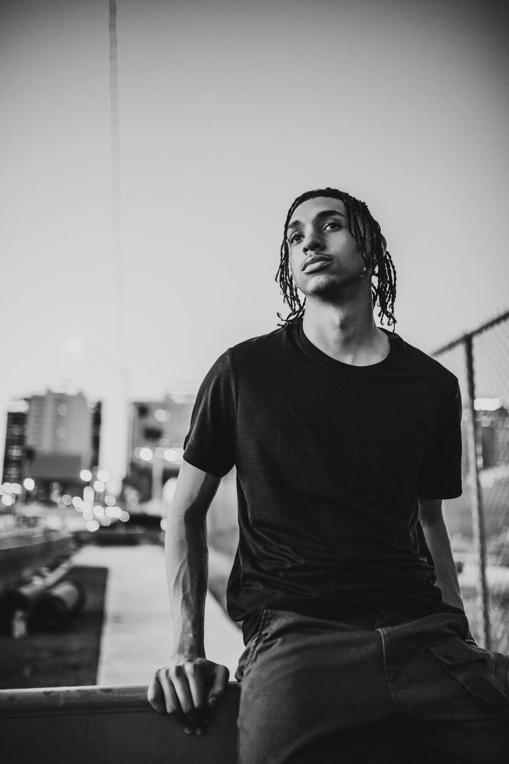 Young man with dreadlocks in black t-shirt poses against urban cityscape in black and white portrait.