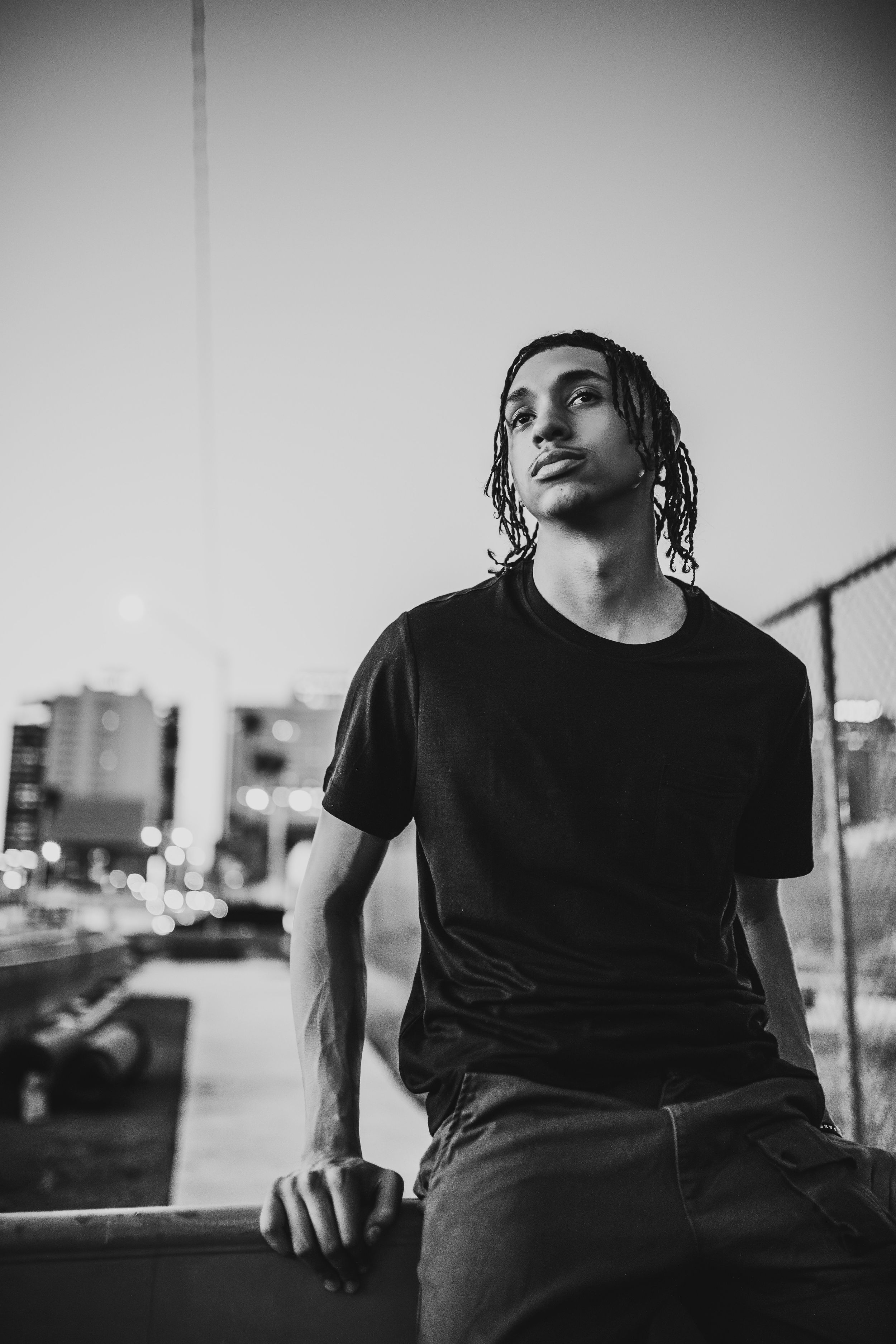 Young man with dreadlocks in black t-shirt poses against urban cityscape in black and white portrait.