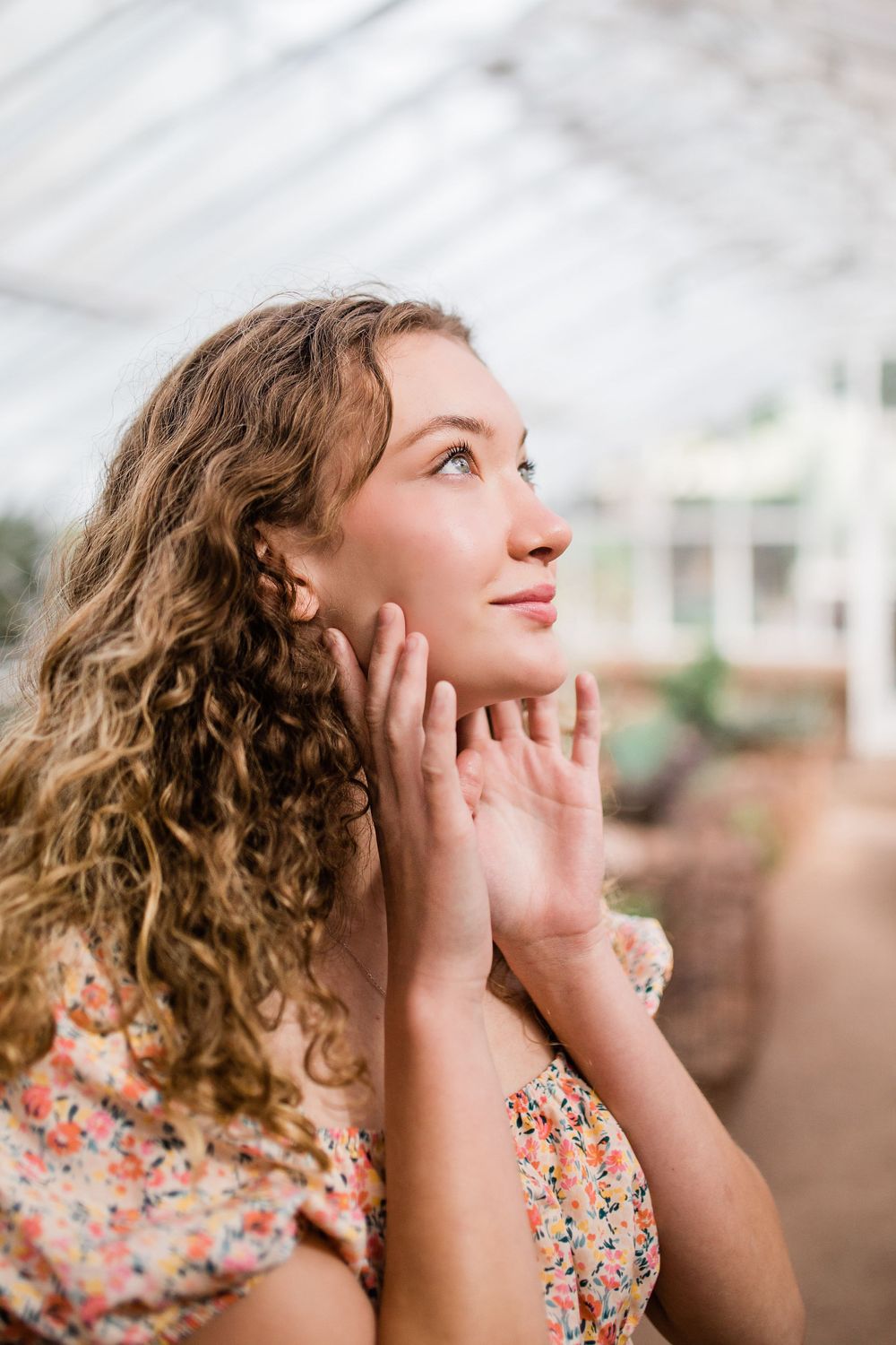 Close-up of curly-haired young woman in floral dress looking upward with hands framing her face.