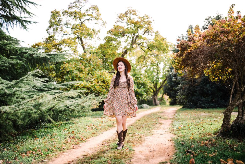 Young woman in floral dress and hat walking on rustic dirt path surrounded by autumn trees.