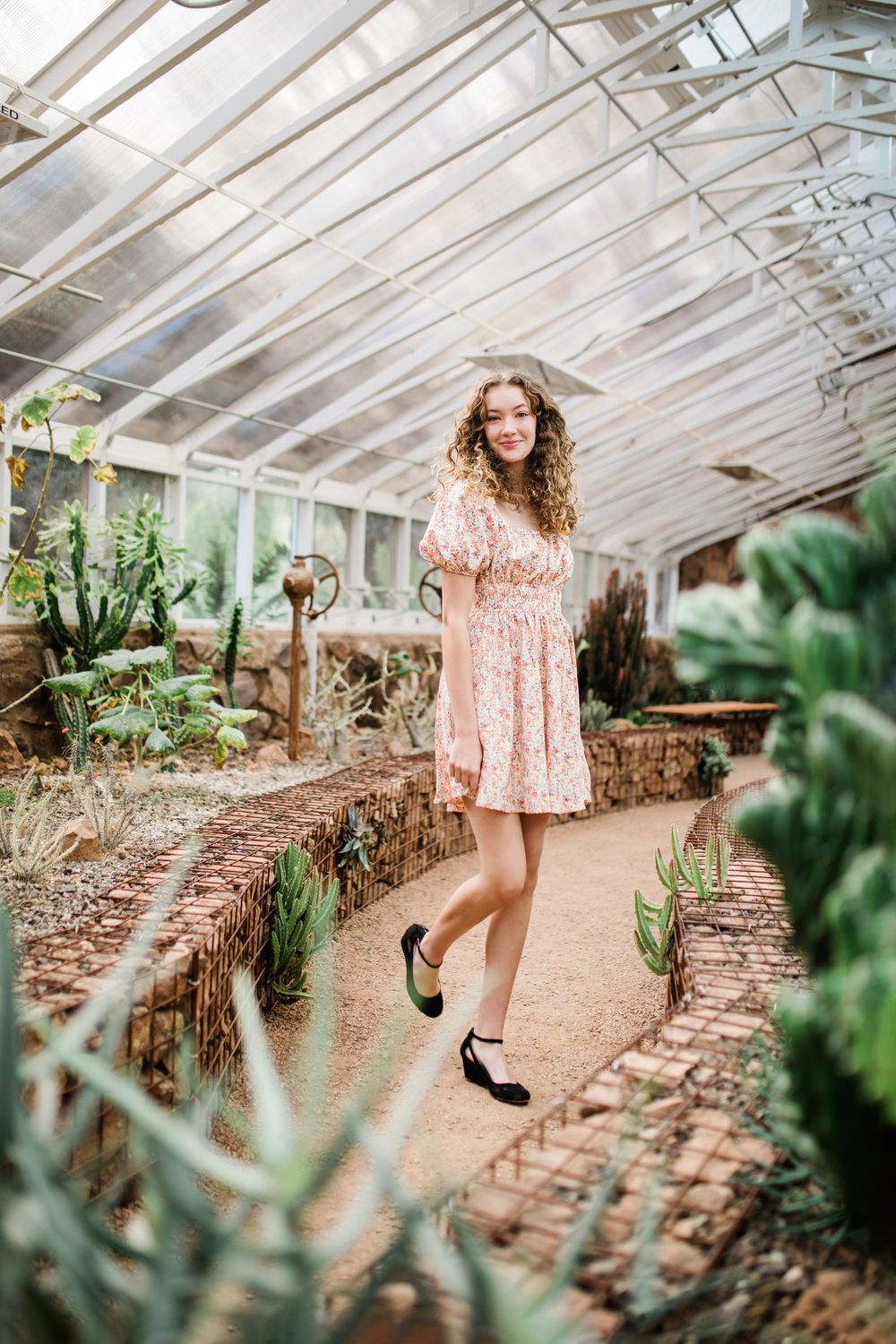Young woman in floral mini dress walking through greenhouse pathway surrounded by cacti.