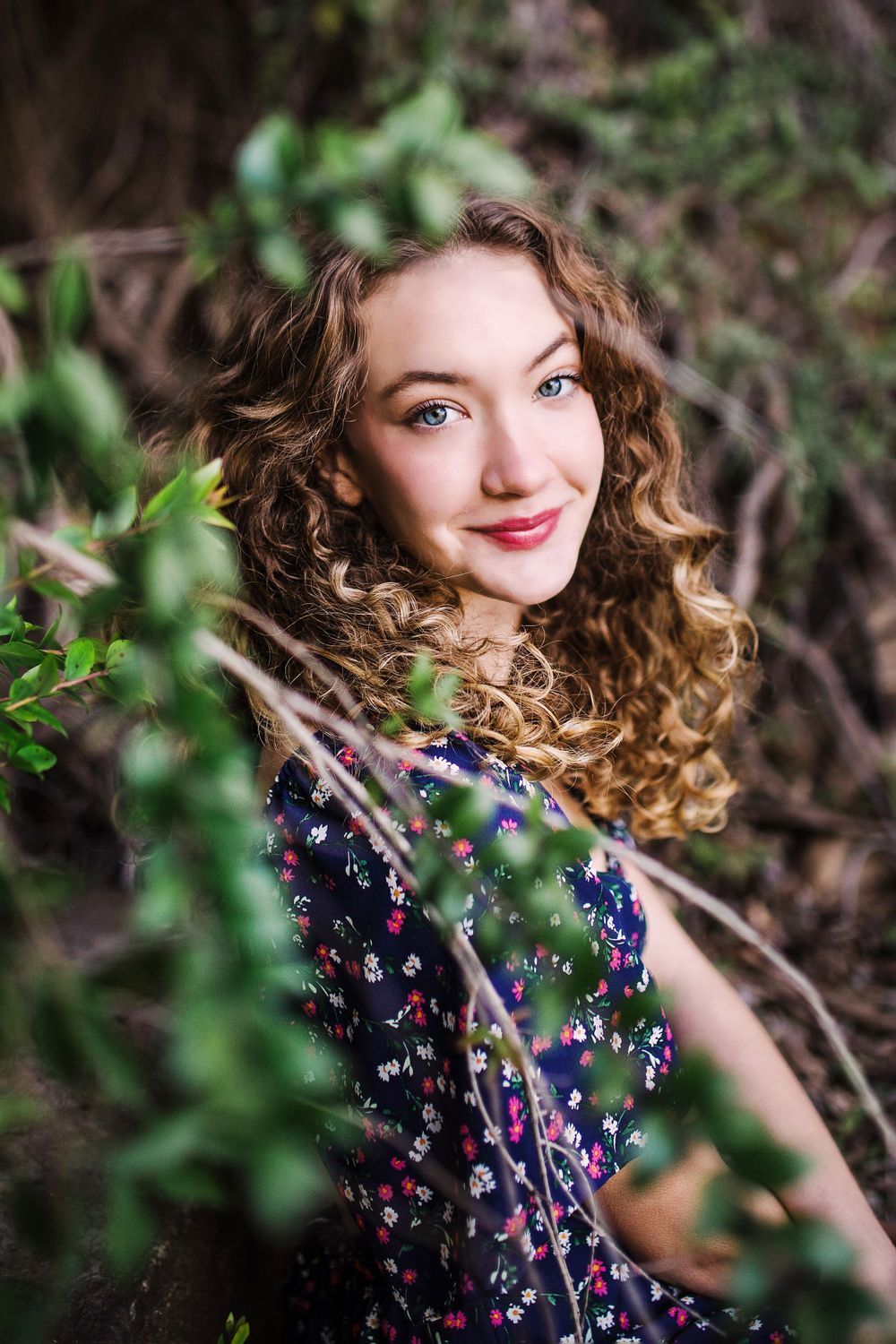 Close-up portrait of a smiling curly-haired woman in a navy floral dress among green branches.