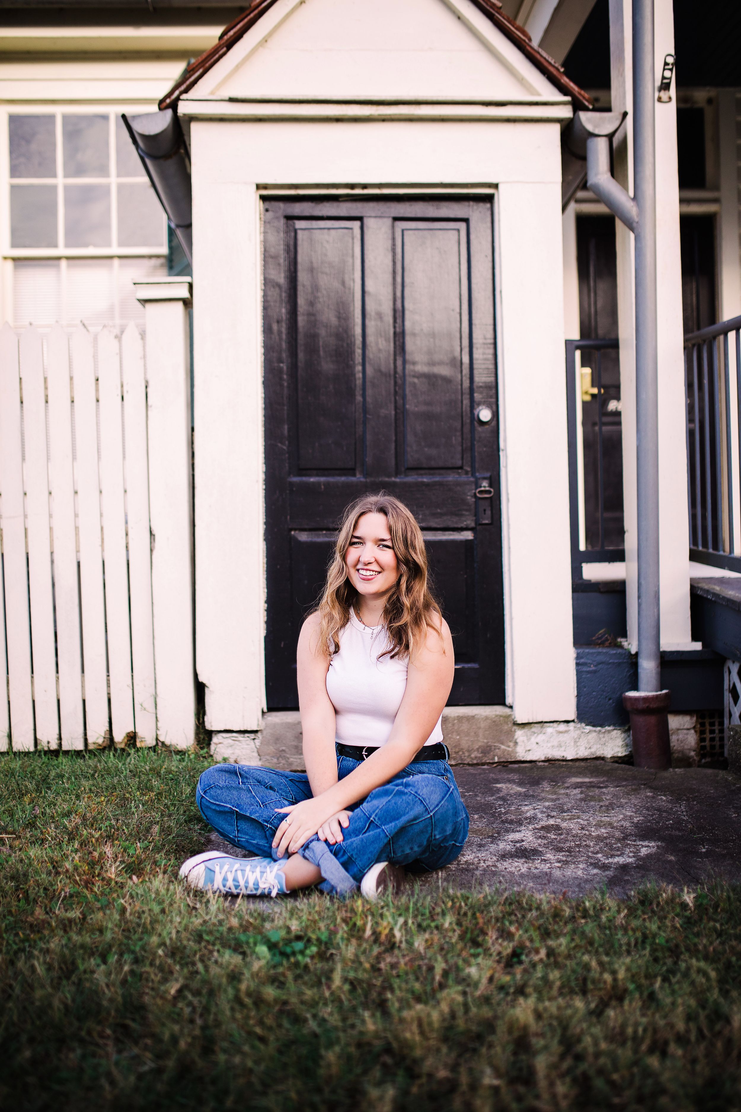 Smiling young woman in white top and jeans sitting cross-legged on grass by a black door.