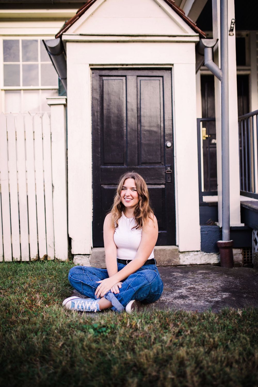 Smiling young woman in white top and jeans sitting cross-legged on grass by a black door.