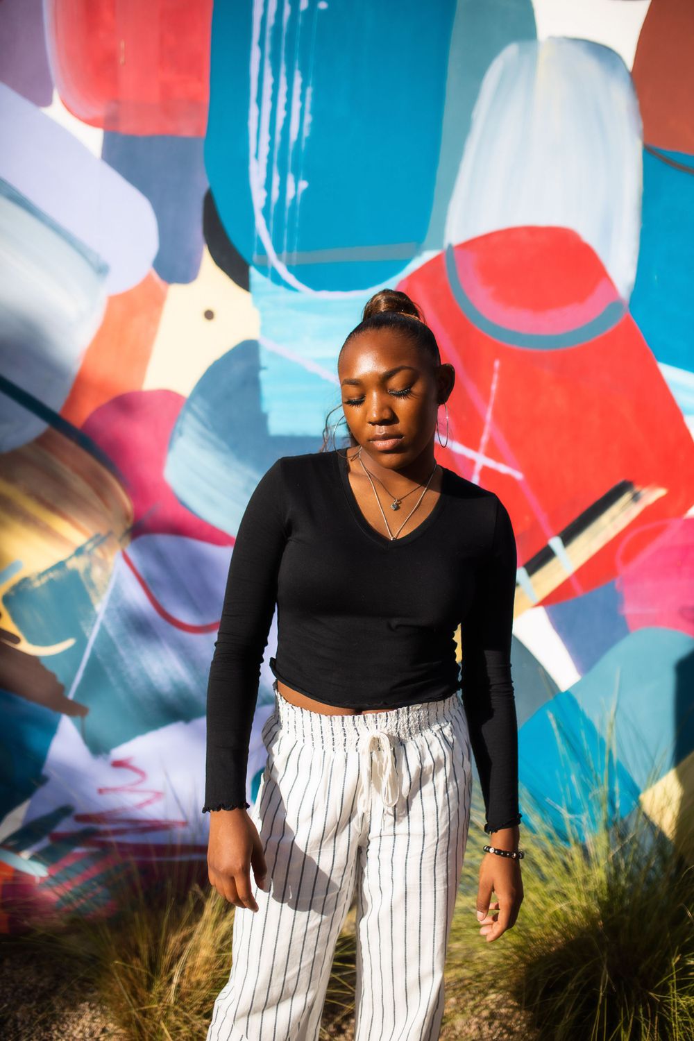 Young Black woman in black crop top and striped pants poses before colorful abstract mural outdoors.