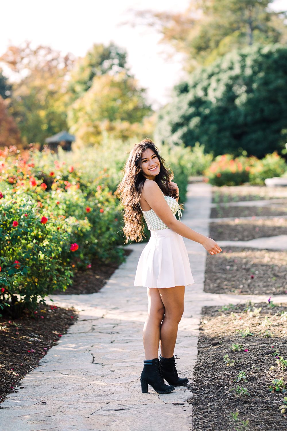 Smiling young woman in white dress and black ankle boots poses on a sunlit rose garden pathway.