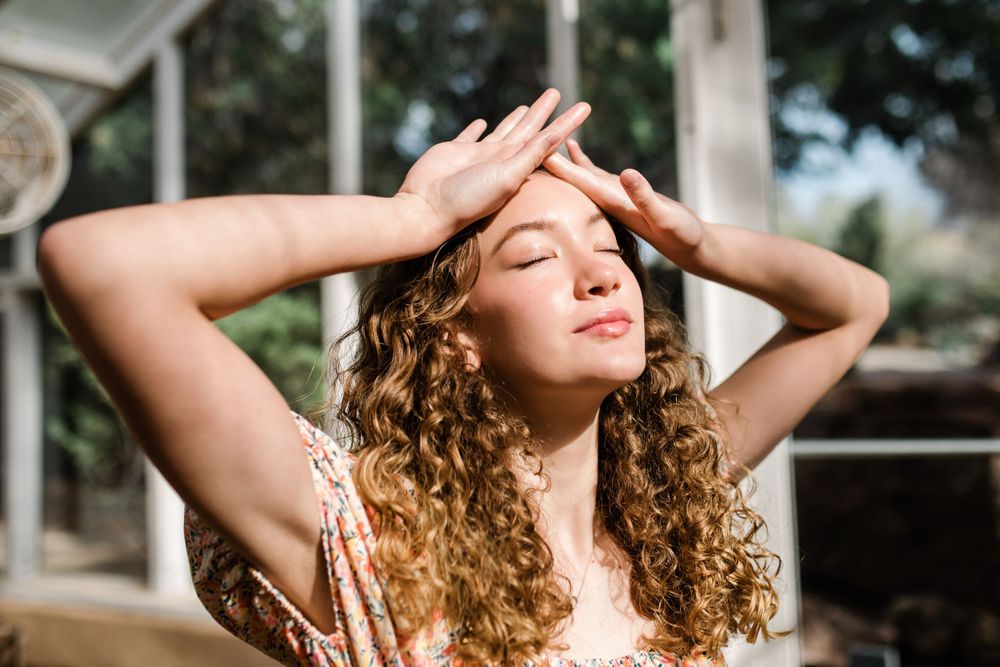Young woman with curly hair posing with hands on head, eyes closed in warm sunlight.