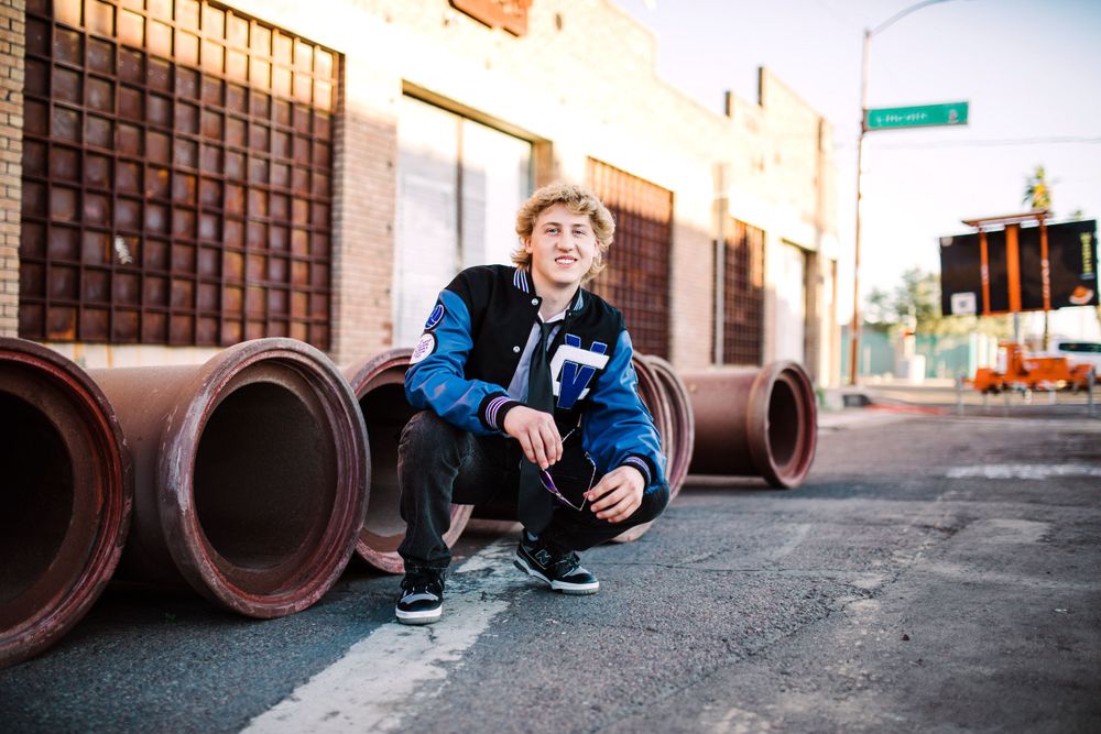 Young man crouching near large industrial pipes in an urban setting wearing a blue jacket.