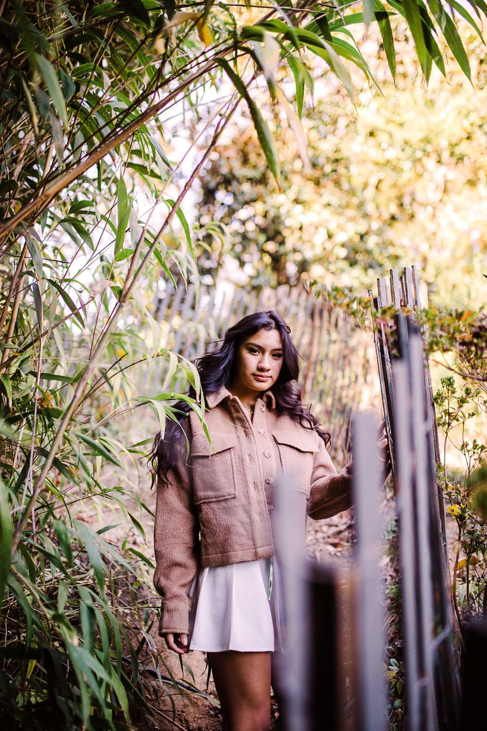 Young woman in brown jacket and white skirt stands among lush bamboo and wooden fence in nature.