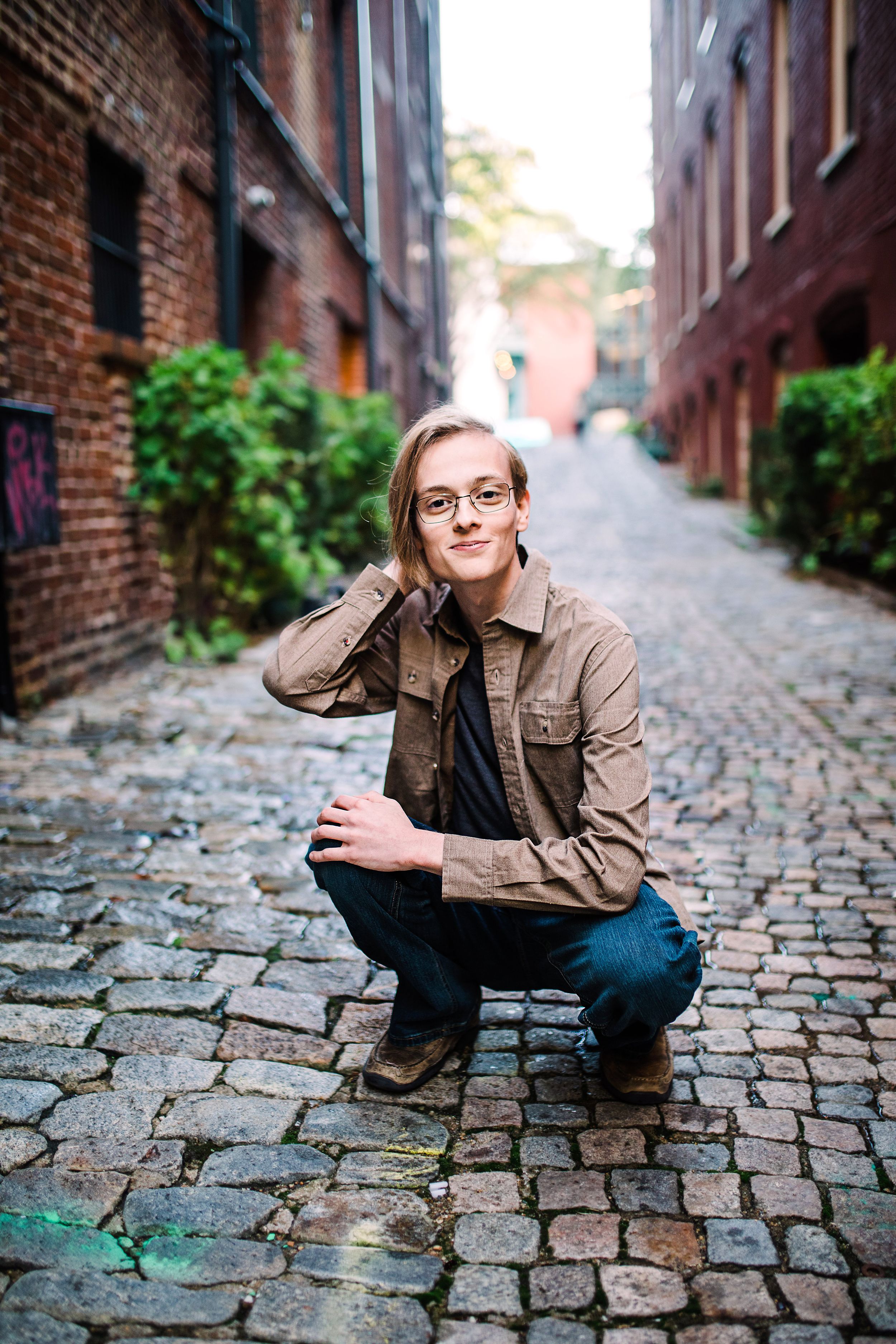 Young man with glasses in brown jacket crouching on cobblestone alley between historic brick buildings.