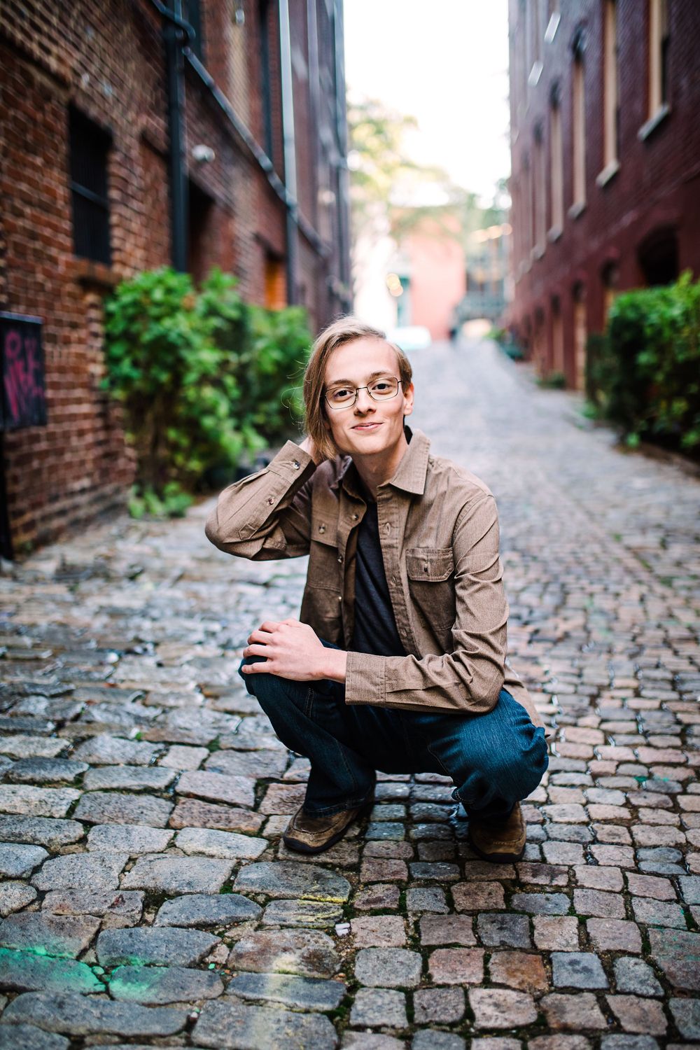 Young man with glasses in brown jacket crouching on cobblestone alley between historic brick buildings.