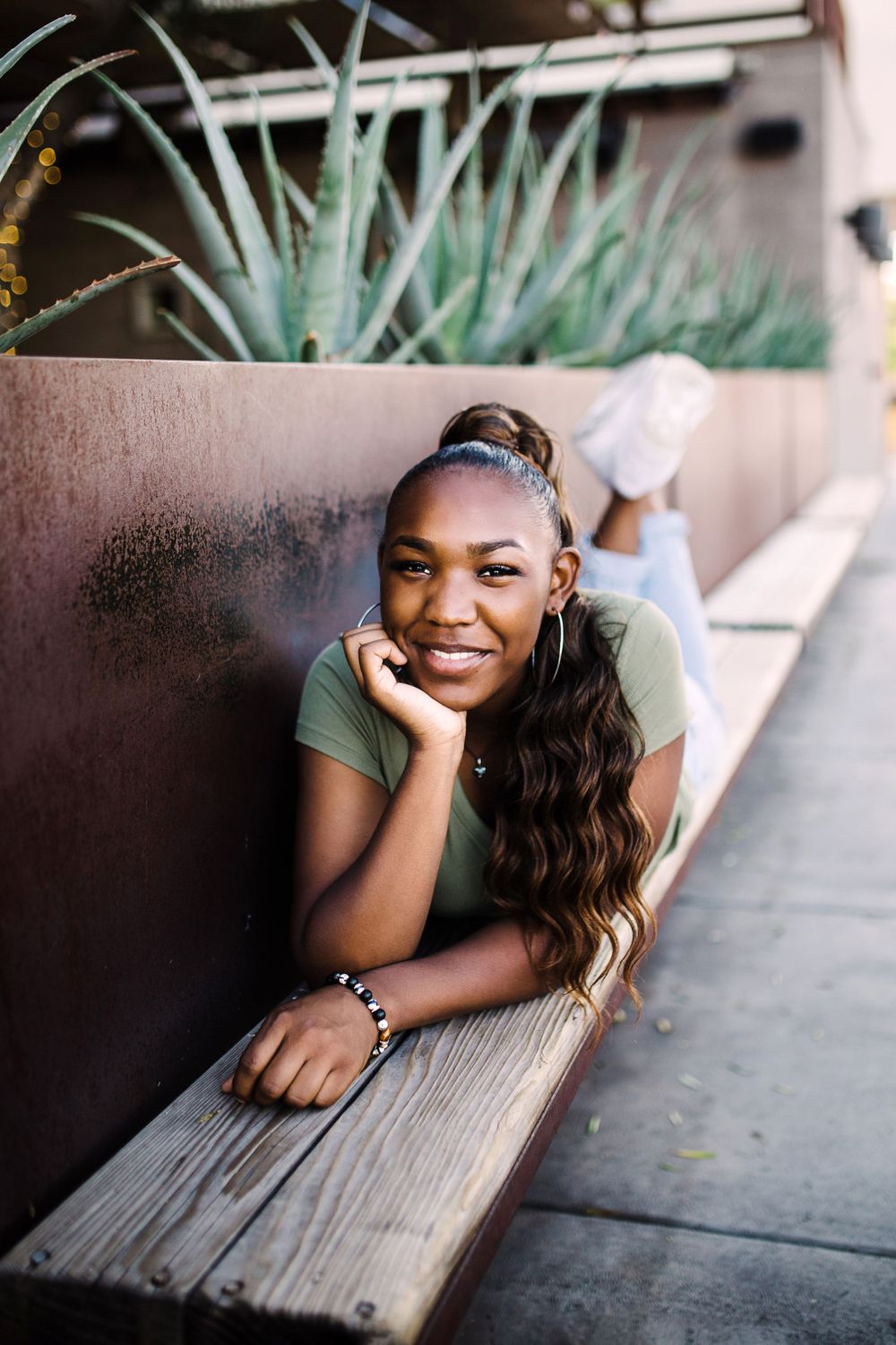 Smiling young Black woman in olive tee lies on wooden ledge beside rustic metal planter with agave.