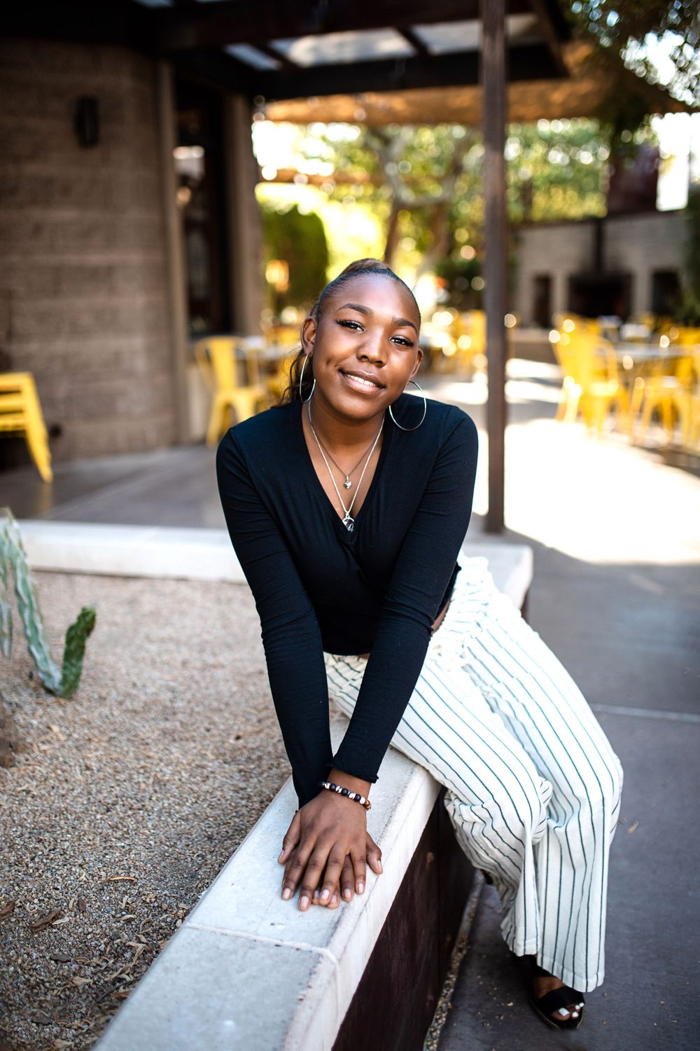 Young Black woman in black top and striped pants leans on ledge at outdoor restaurant patio.