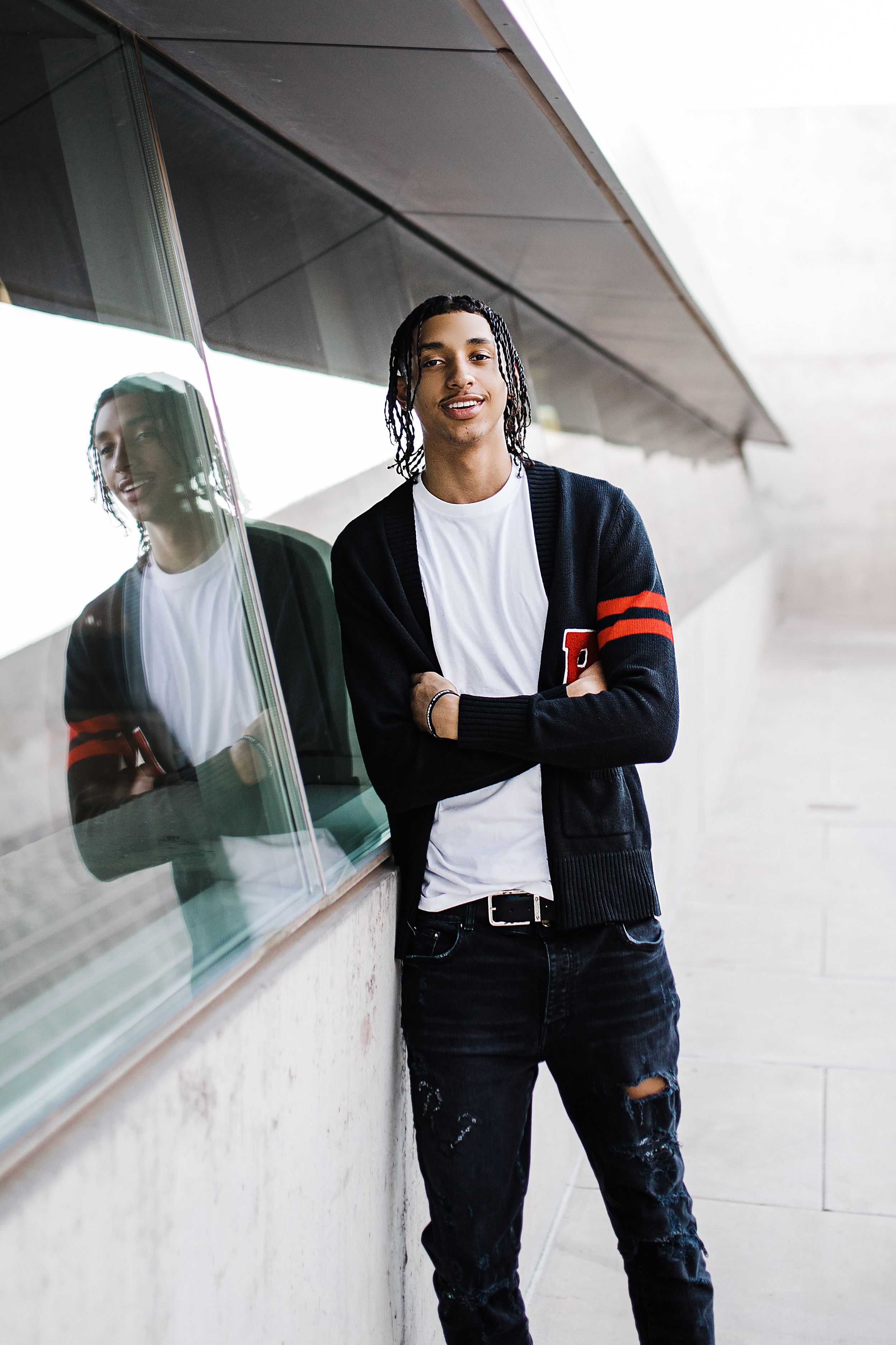 Young man with braids in black varsity cardigan leans against glass wall with reflection visible.