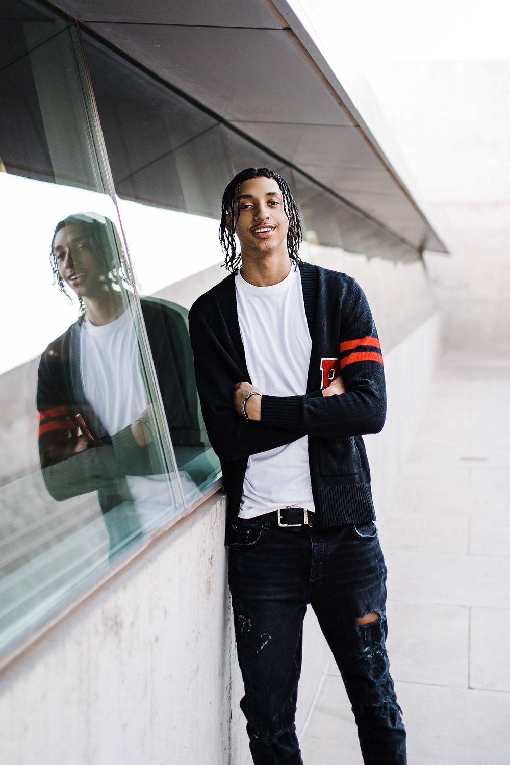 Young man with braids in black varsity cardigan leans against glass wall with reflection visible.
