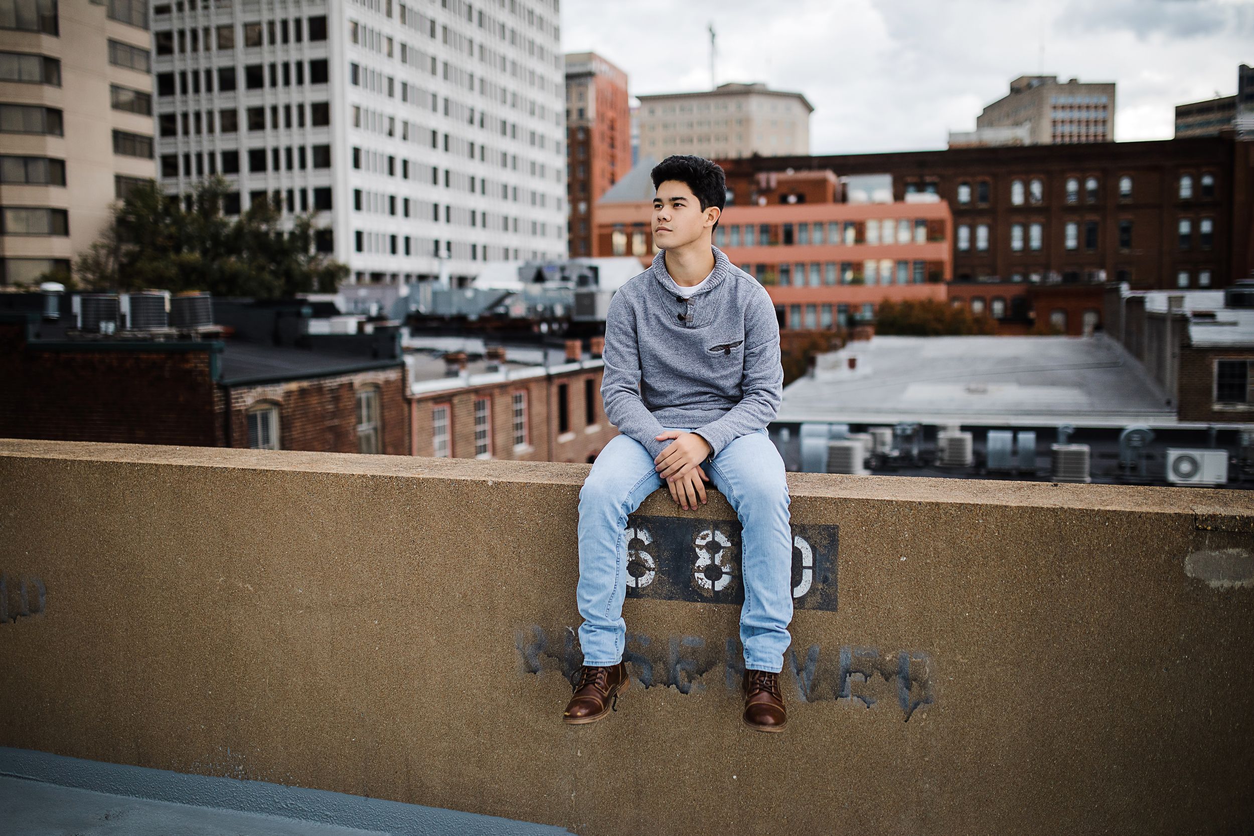 Young man sitting on urban rooftop ledge with city buildings in background.