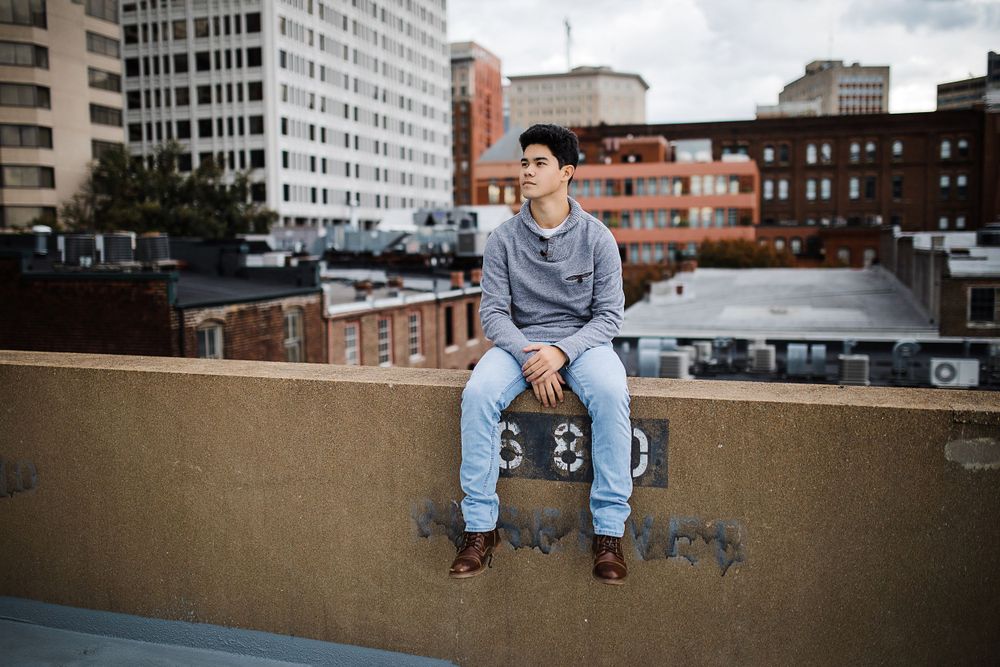 Young man sitting on urban rooftop ledge with city buildings in background.