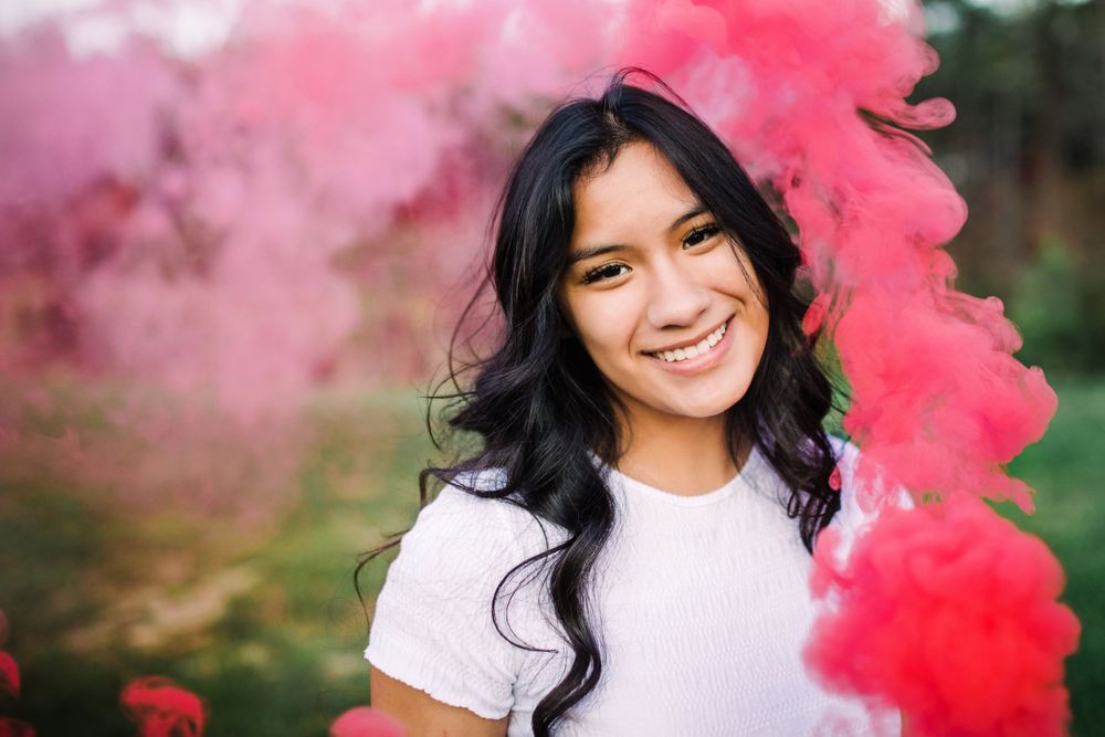 Smiling young woman with dark curly hair surrounded by pink and red smoke outdoors.