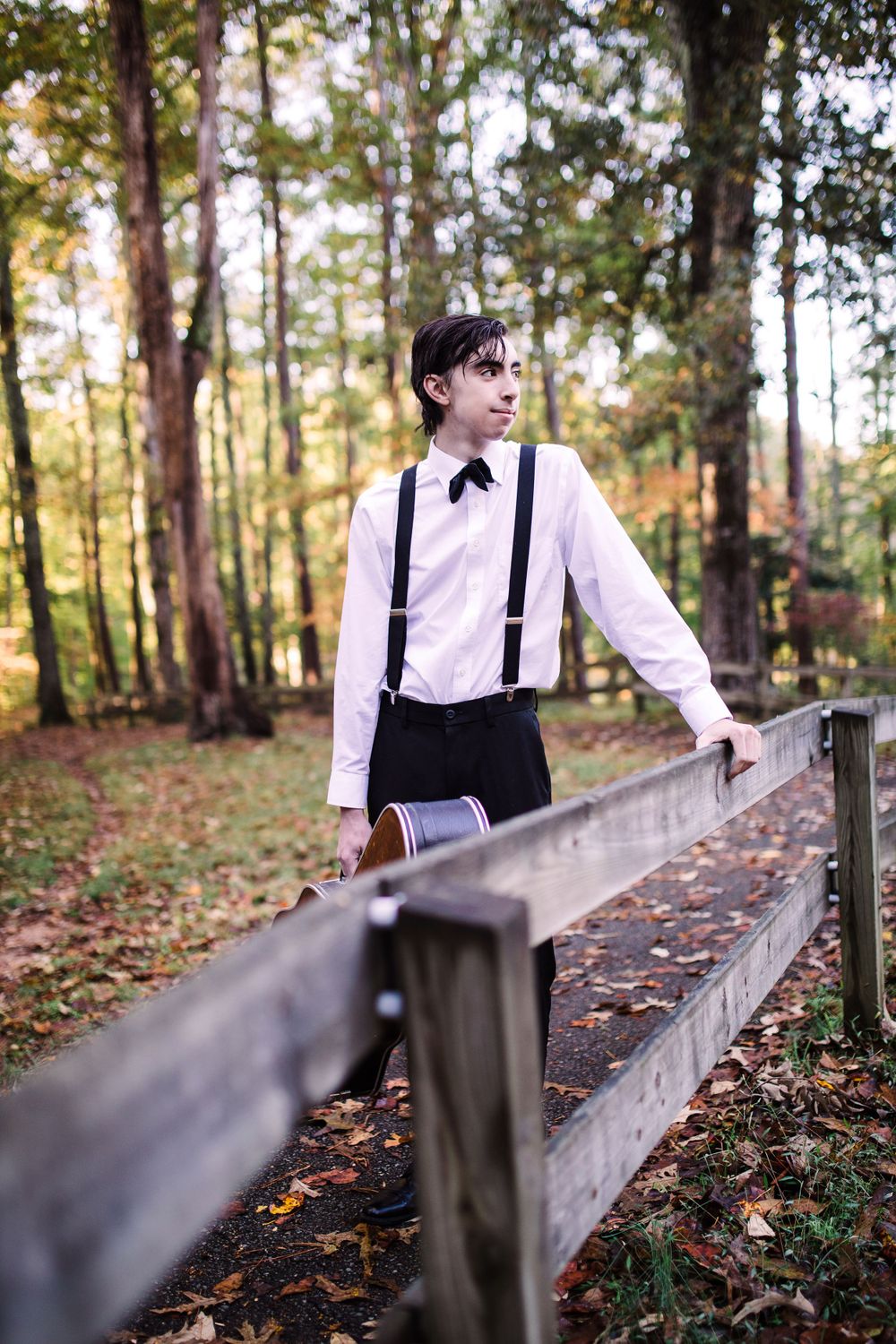 Teen boy in white shirt, suspenders and bow tie leaning on wooden fence in autumn forest setting.
