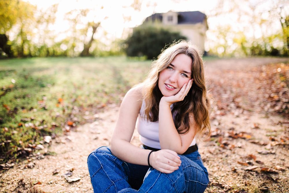 Young woman in white tank and jeans sitting outdoors on a dirt path with warm autumn sunlight.
