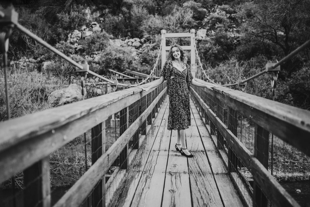 Black and white photo of a curly-haired woman in a floral dress standing on a wooden suspension bridge.
