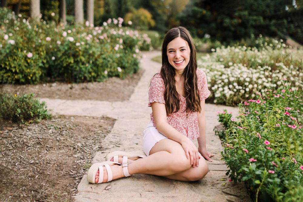 Happy young woman in pink floral blouse and white shorts sitting on garden path surrounded by flowers.