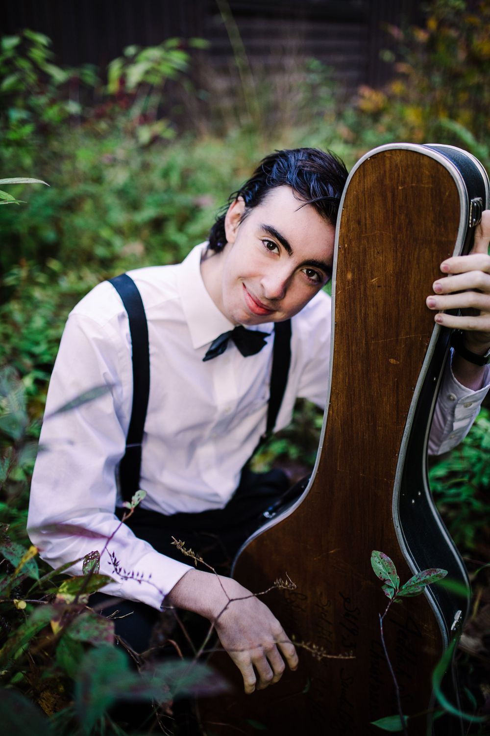 Young man in suspenders and bow tie leaning on vintage guitar case surrounded by green foliage.